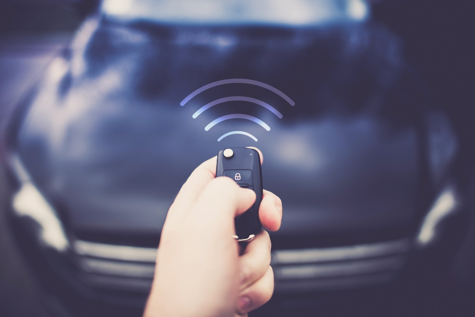 A hand presses a car key fob, emitting a signal toward a vehicle in the background. This illustrates remote locking or unlocking features.